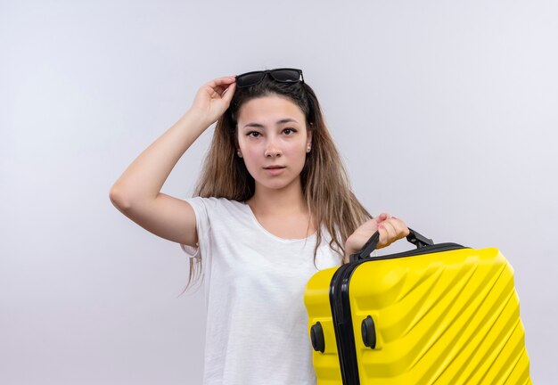 Молодая путешественница в белой футболке держит чемодан с озадаченным видом, стоя у белой стены