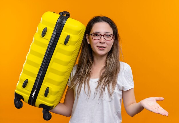 Young traveler woman in white t-shirt  holding suitcase looking uncertain and confused standing over orange wall