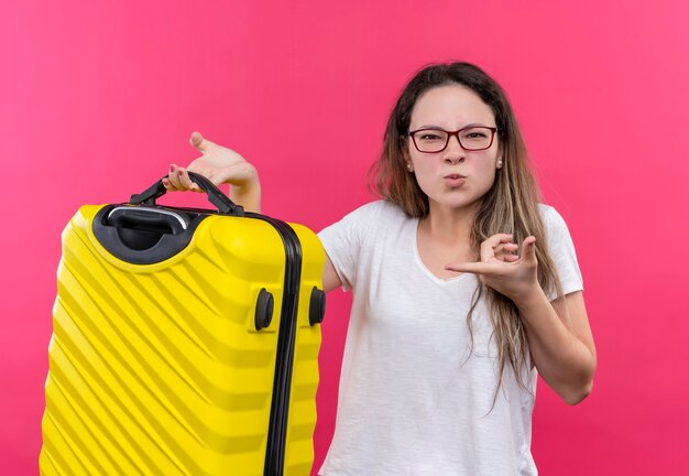 ピンクの壁の上に立ってイライラして見える人差し指でそれを指している旅行スーツケースを保持している白いTシャツの若い旅行者の女性