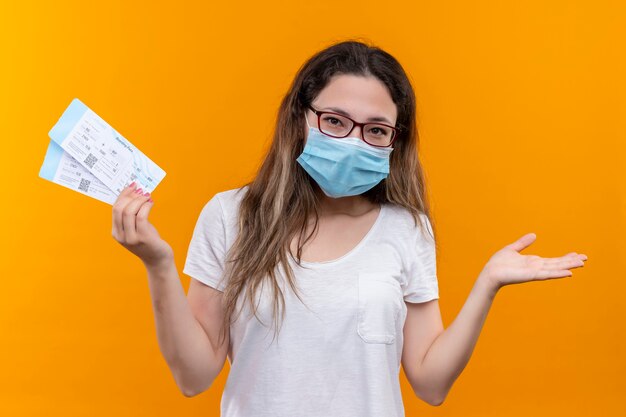 Young traveler woman in white t-shirt wearing protective facial mask holding air tickets smiling raising hand standing over orange wall