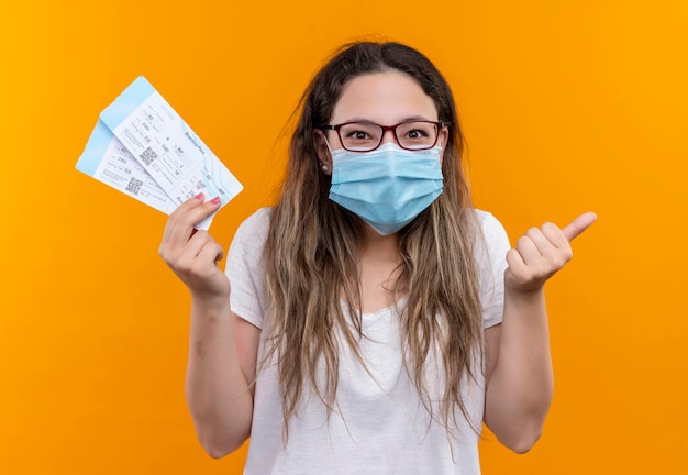 Young traveler woman in white t-shirt wearing protective facial mask holding air tickets smiling showing thumbs up standing over orange wall