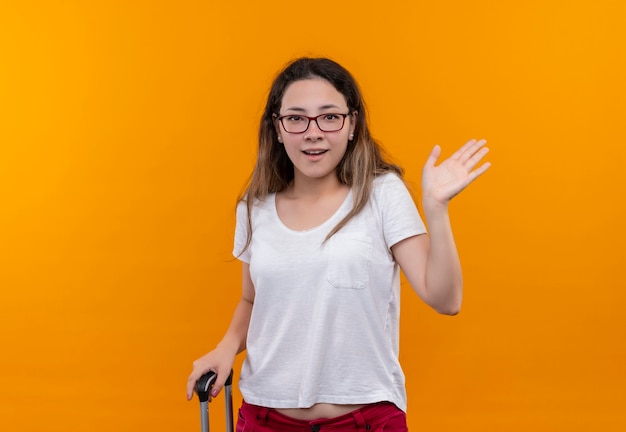 Free photo young traveler woman in white t-shirt with suitcase surprised and happy waving with hand standing over orange wall