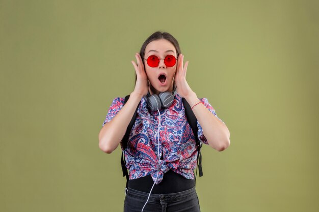 Молодой путешественник женщина с рюкзаком и наушниками, носить красные очки шокирован стоя с широко открытым ртом трогательно лицо руками над зеленой стеной