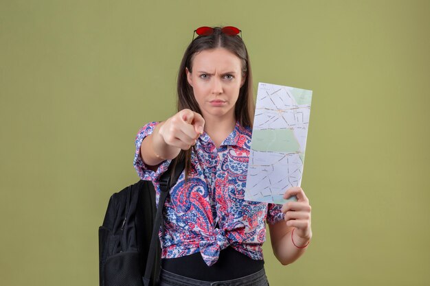 Young traveler woman with red sunglasses on head and with backpack holding map displeased pointing to camera with finger with angry expression over green wall