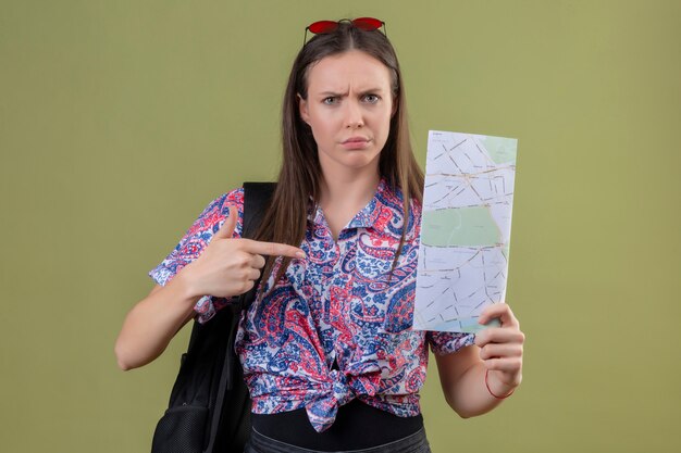Молодой путешественник женщина с красными очками на голове и с рюкзаком, держа карту, указывая указательным пальцем к нему с хмурым лицом над зеленой стеной