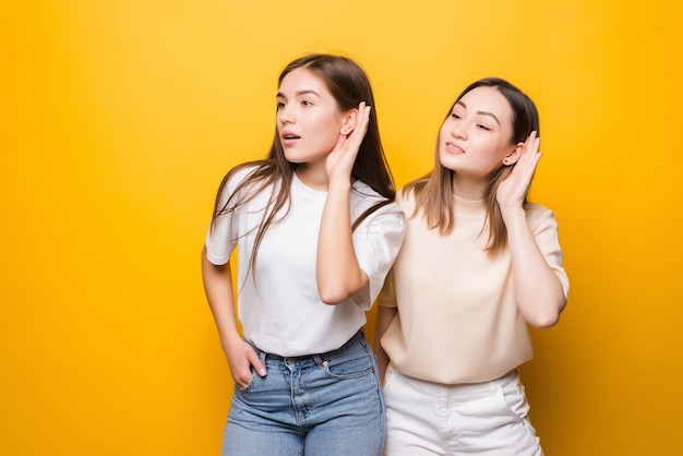 黄色の壁に隔離された耳に手を置くことによって何かを聞いている若い2人の女性
