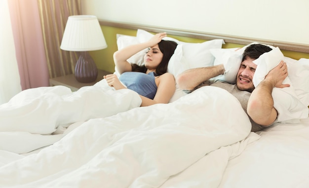 Young unhappy couple covering ears and having headache while sleeping on bed