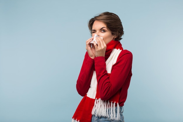 Young unhealthy lady with dark short hair in red sweater and scarf blowing nose with white napkin over blue background isolated