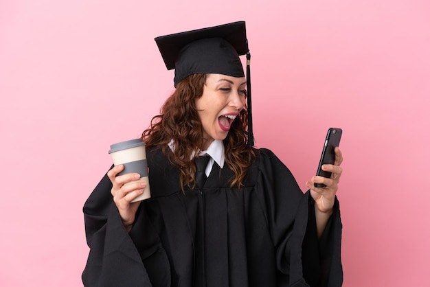 Молодая женщина-выпускница университета изолирована на розовом фоне, держа кофе на вынос и мобильный