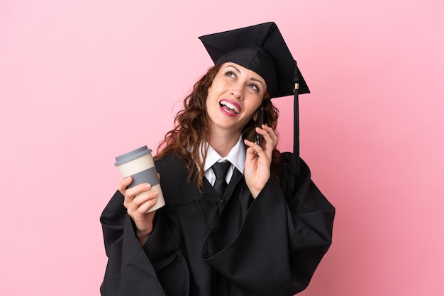 Молодая выпускница университета, изолированная на розовом фоне, держит кофе на вынос и мобильный телефон