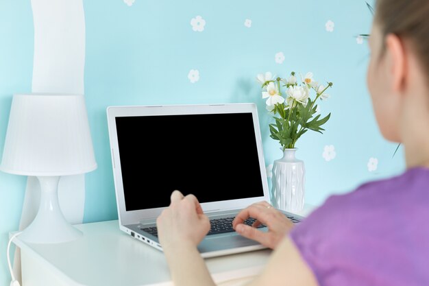 Young unrecognizable woman freelancer works remotely at home, sits on cozy domestic blue interior in front of opened modern laptop computer with blank copy screen for your promotional text