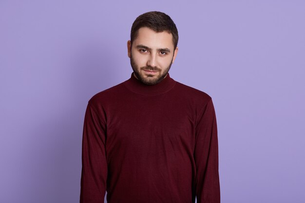Young unshaved man wearing burgundy sweater posing against purple background with serious facial expression