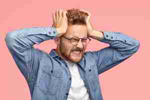 Foto gratuita giovane uomo stressante con la barba lunga soffre di mal di testa, tiene le mani sulle tempie, stringe i denti, ha un dolore terribile, chiude gli occhi