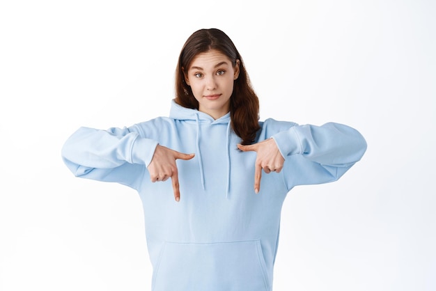 Young unsure girl in hoodie pointing fingers down raising eyebrows with doubtful expression having hesitations standing against white background