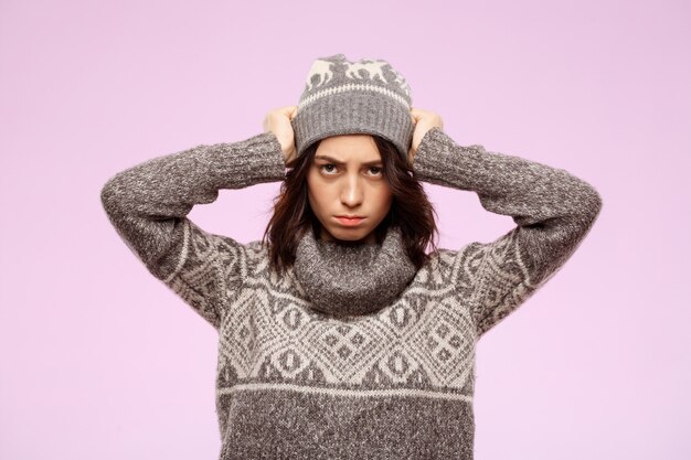 Young upset beautiful brunette girl in hat and sweater  over light wall
