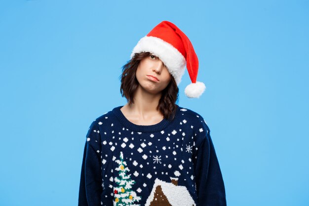 Young upset beautiful brunette girl in knitted sweater and christmas hat over blue wall