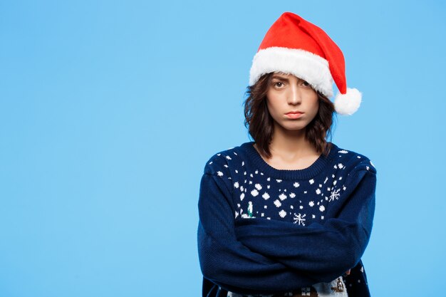 Young upset beautiful brunette girl in knitted sweater and christmas hat over blue wall