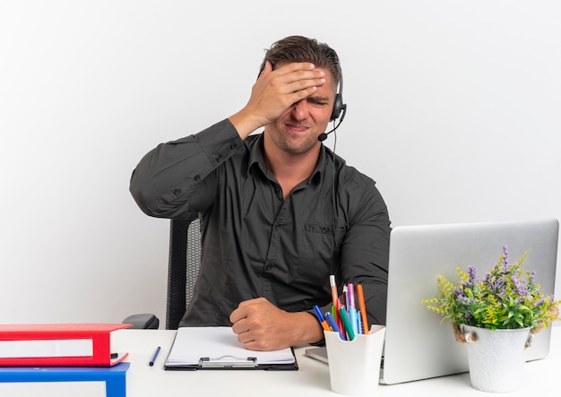 Молодой расстроенный белокурый человек офисного работника в наушниках сидит за столом с офисными инструментами, используя ноутбук, кладет руку на голову, изолированную на белом фоне с копией пространства