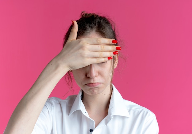 Young upset blonde russian girl hides eyes with hand isolated on pink space with copy space