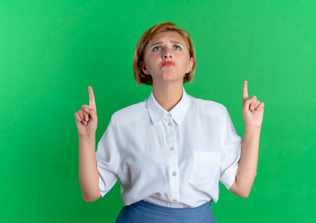 Young upset blonde russian girl points up isolated on green background with copy space