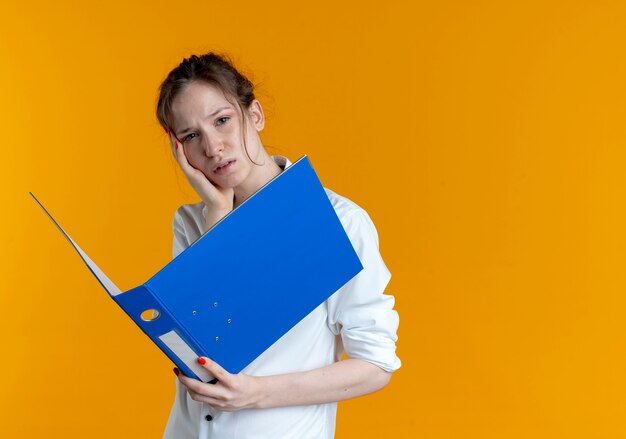 Young upset blonde russian girl puts hand on chin holding file folder isolated on orange space with copy space