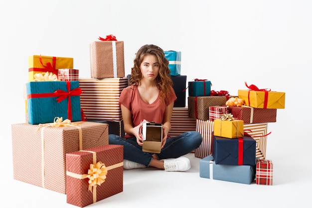 Young upset curly woman sitting on floor among gift boxes showing one she opened is empty