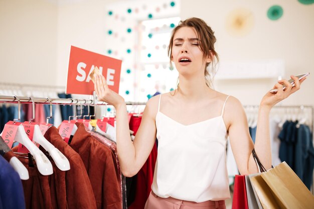 Young upset girl in white top standing near sale clothes rack with credit card and colorful shopping bags in hands in boutique. Portrait of depress lady with cellphone in hand in clothes store