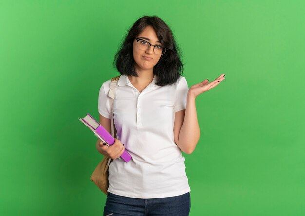 Молодая расстроенная симпатичная кавказская школьница в очках и задней сумке держит руку прямо, держа книги на зеленом с копией пространства