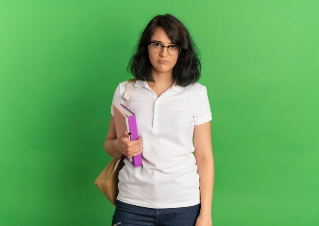 Young upset pretty caucasian schoolgirl wearing glasses and back bag stands holding books on green  with copy space