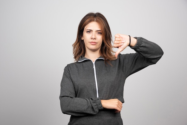 Young upset woman showing thumbs down on a gray wall.