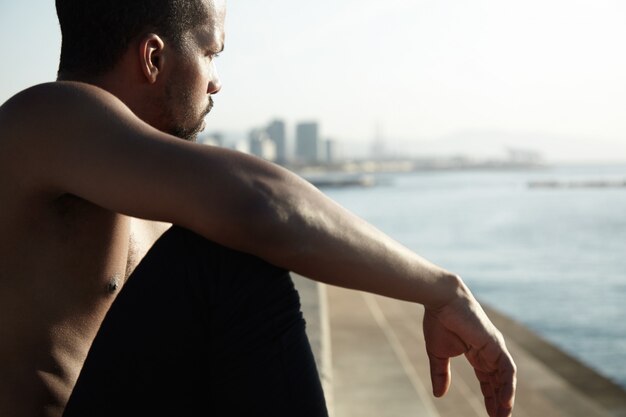 Young vagabond sitting on the riverwalk with stretched arm resting on his knee. Black-skinned man is thinking about his life in big city and watching waves of water, relaxing under the sunlight.