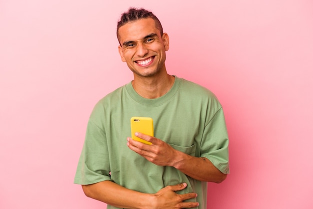 Young venezuelan man holding a mobile phone isolated on pink background laughing and having fun.