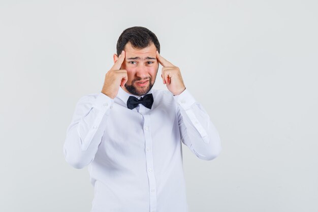 Young waiter holding fingers on temples and having headache in white shirt front view.