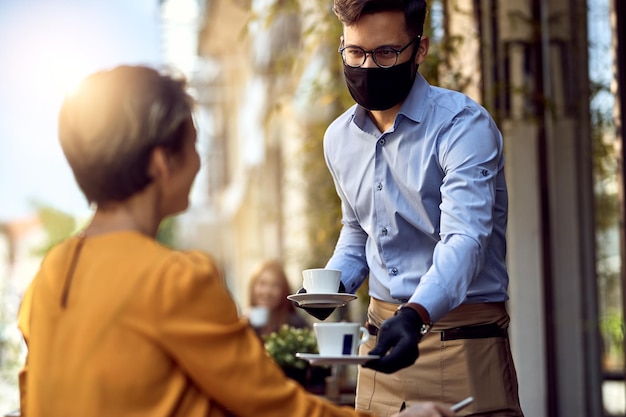 Young waiter wearing protective face mask while serving coffee to a customer in a cafe after reopening