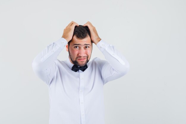 Young waiter in white shirt clasping head in hands and looking helpless , front view.