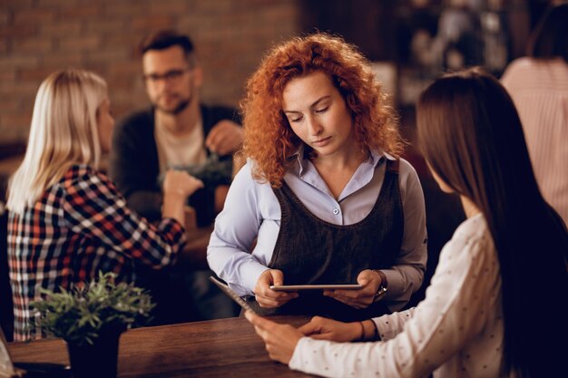 Young waitress helping female guest to choose something from the menu while taking an order in a pub