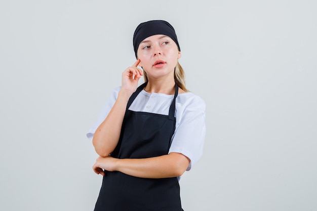 Young waitress holding finger on cheek in uniform and apron and looking pensive