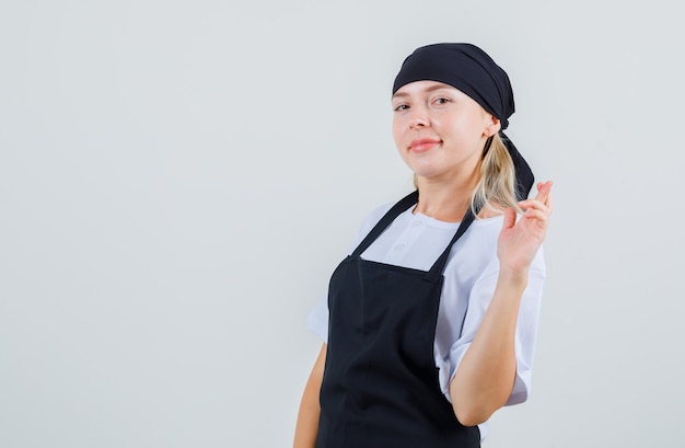 Young waitress holding fingers crossed in uniform and apron and looking merry