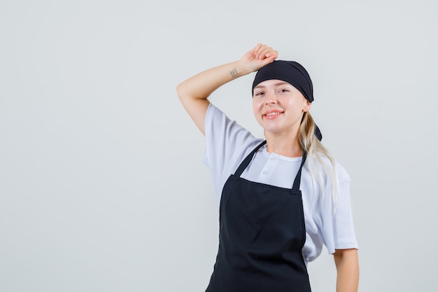 Free photo young waitress holding fist on head in uniform and apron and looking cheery