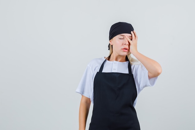 Free photo young waitress holding hand on face in uniform and apron and looking forgetful