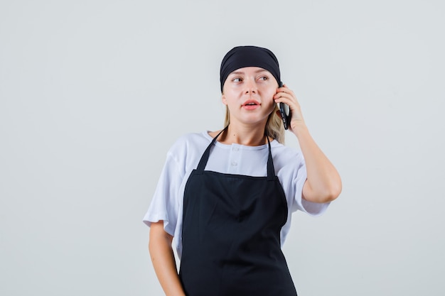 Бесплатное фото Молодая официантка в униформе и фартуке смотрит в сторону во время разговора по мобильному телефону
