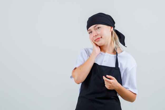 Young waitress leaning cheek on raised palm in uniform and apron and looking cheerful