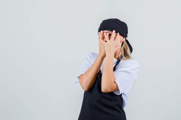 Young waitress looking through fingers in uniform and apron and looking scared