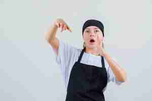 Free photo young waitress pointing at her raised palm in uniform and apron and looking surprised