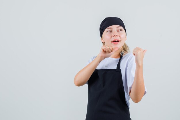 Young waitress pointing thumbs back in uniform and apron and looking cheery