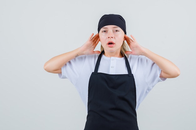 Giovane cameriera che appoggia le dita sulle orecchie in uniforme e grembiule e sembra infastidita