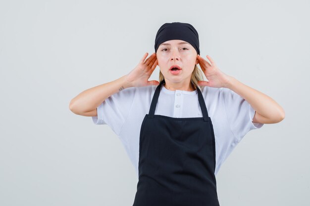 Young waitress propping fingers on ears in uniform and apron and looking annoyed