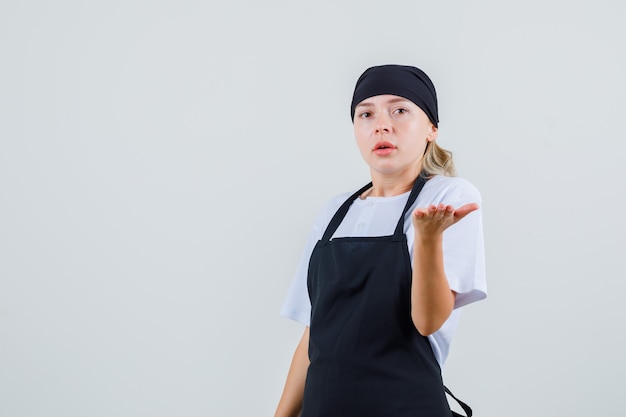 Young waitress raising palm in questioning manner in uniform and apron