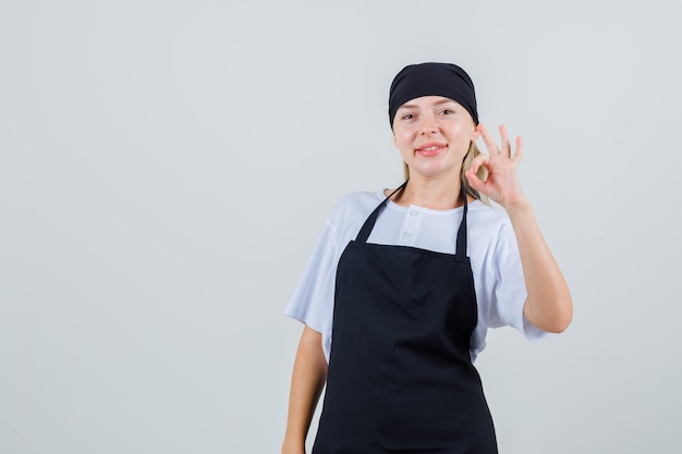 Young waitress showing ok gesture in uniform and apron and looking cheerful