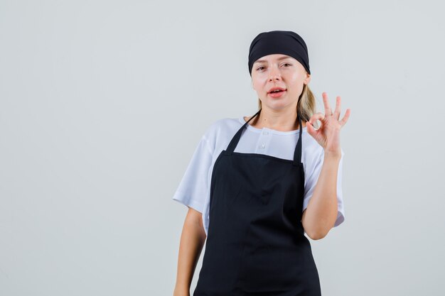 Young waitress showing ok gesture in uniform and apron and looking confident